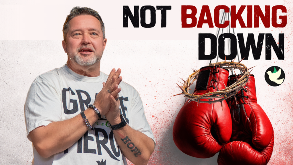 Man with hands clasped in front of a gray T‑shirt, beside red boxing gloves wrapped in a thorn crown, with the text 'NOT BACKING DOWN' above.